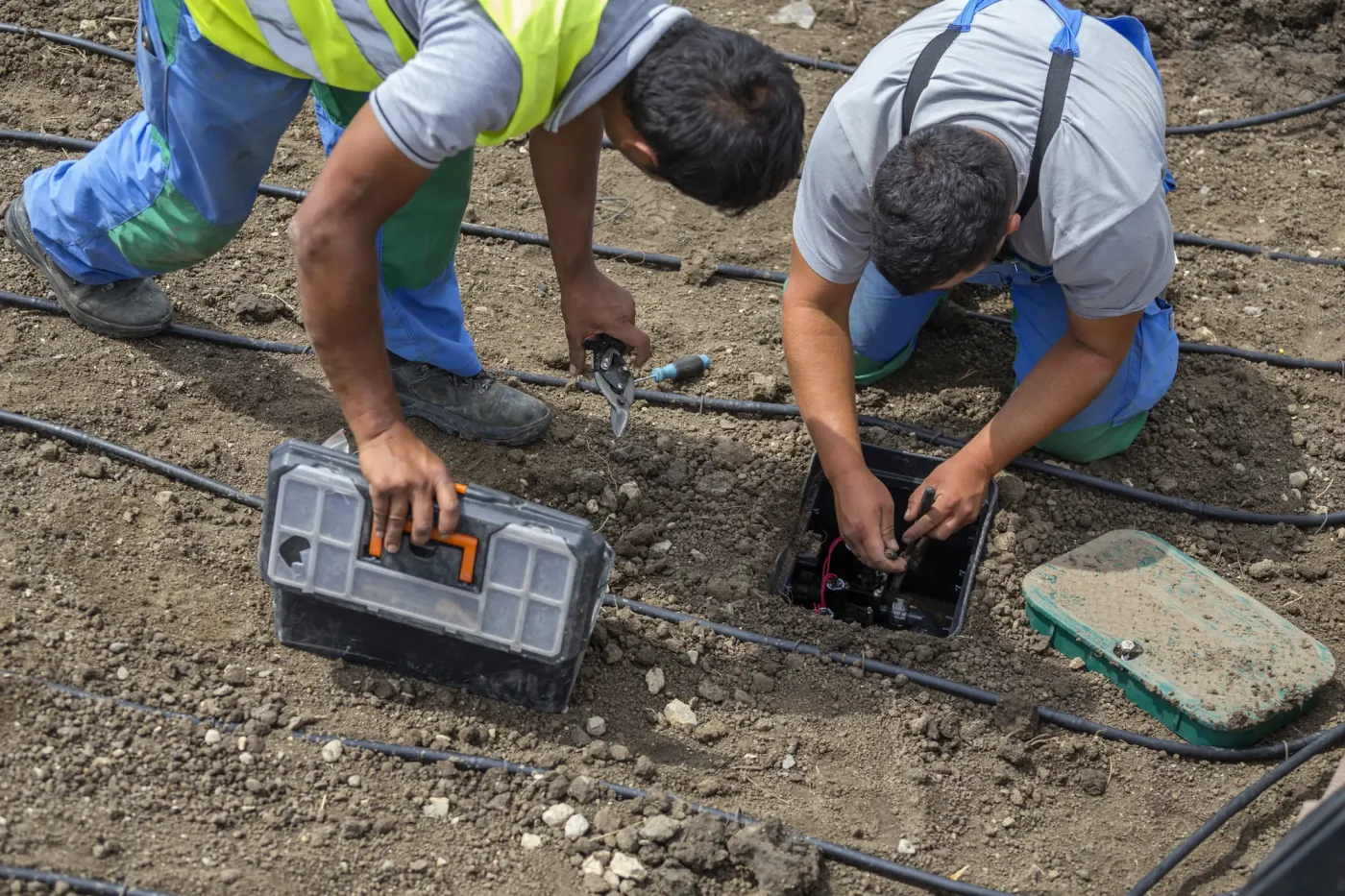 Two workers repair a Palm Beach County, FL sprinkler system wiring on soil using tools and a toolbox.