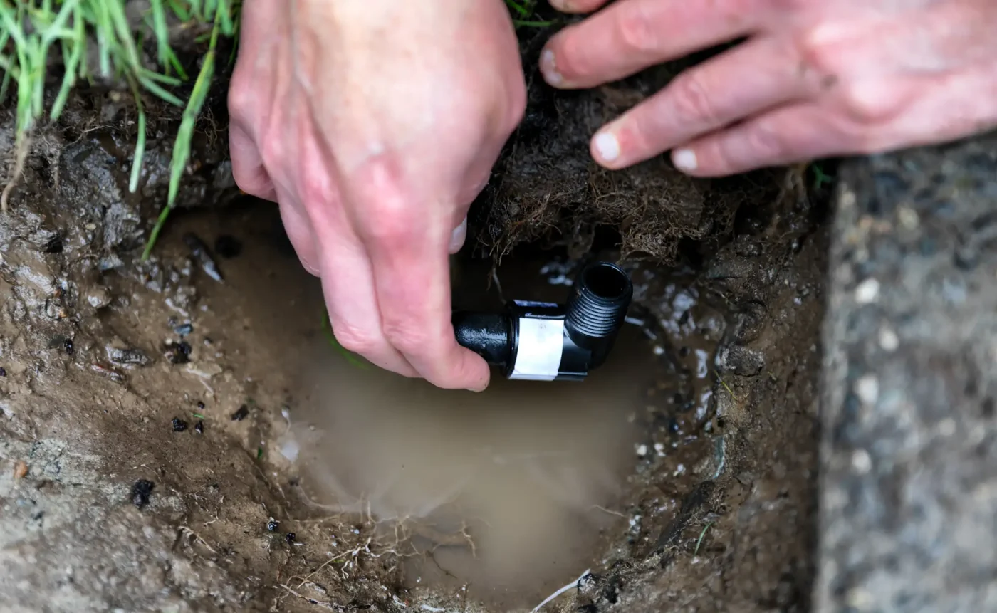 Hands fixing a black elbow joint pipe in muddy water, showing expert sprinkler repair work in Palm Beach County, FL.