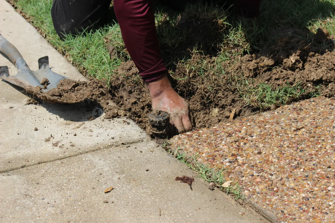 A person digs in soil next to a Palm Beach County, FL sidewalk, likely performing sprinkler repair.